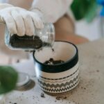 A person wearing white gloves is pouring soil from a glass jar into a decorative ceramic pot, with scattered soil on the table, depicting the process of preparing soil for planting.