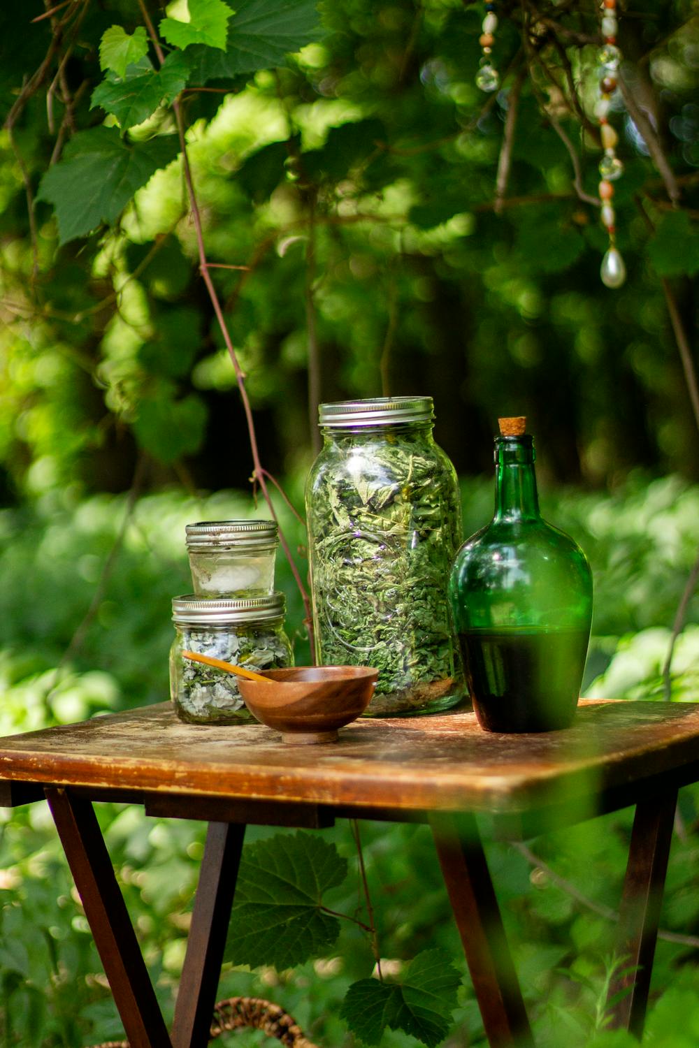Glass jars filled with dried herbs and a green glass bottle placed on a rustic wooden table in a lush green forest setting.